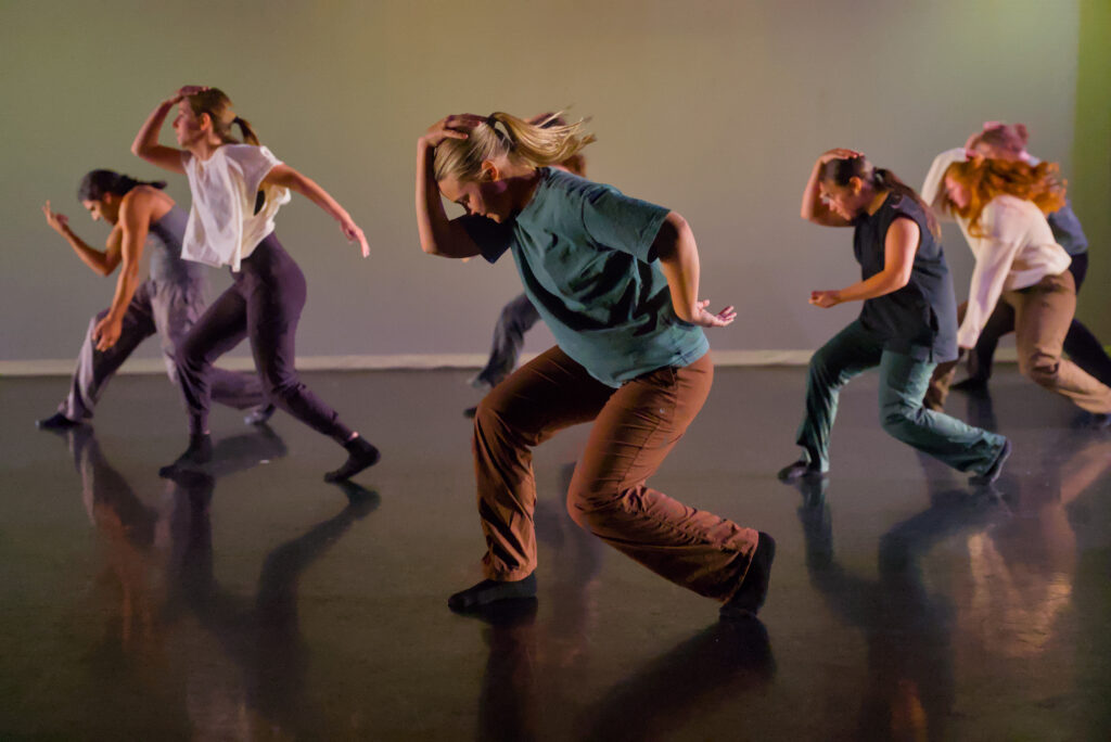 Eight college dance students perform in a black box theater in front of a light background. Each is crouched down and turned to the right, with their right hands on their heads.