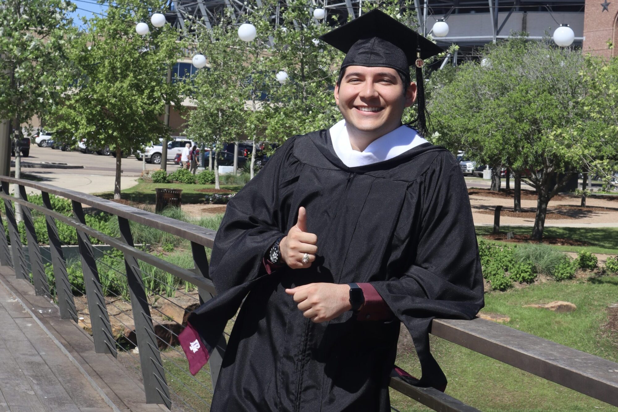 A college graduate student wearing a traditional graduation cap and gown stands on a bridge over a pond, giving the thumbs-up sign.