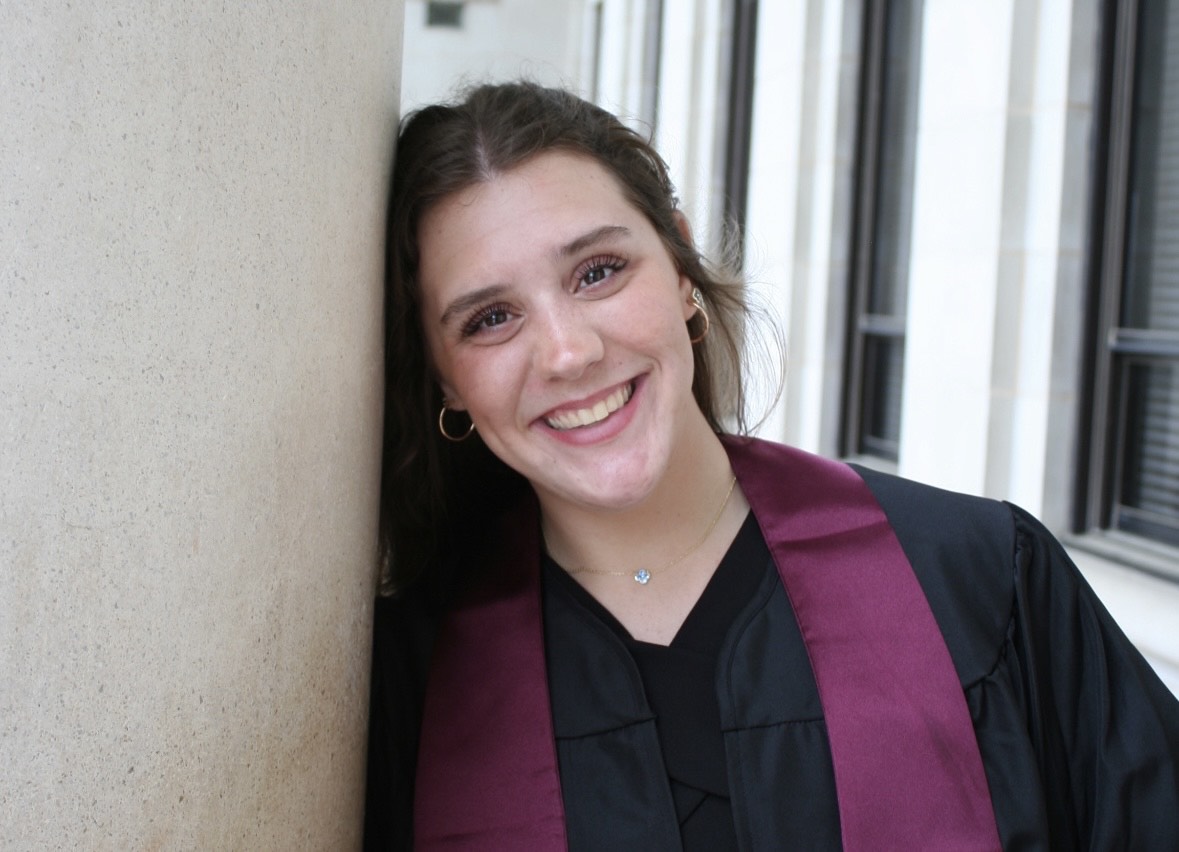 A college student wears a traditional graduation gown and a maroon sash as she smiles for a portrait.