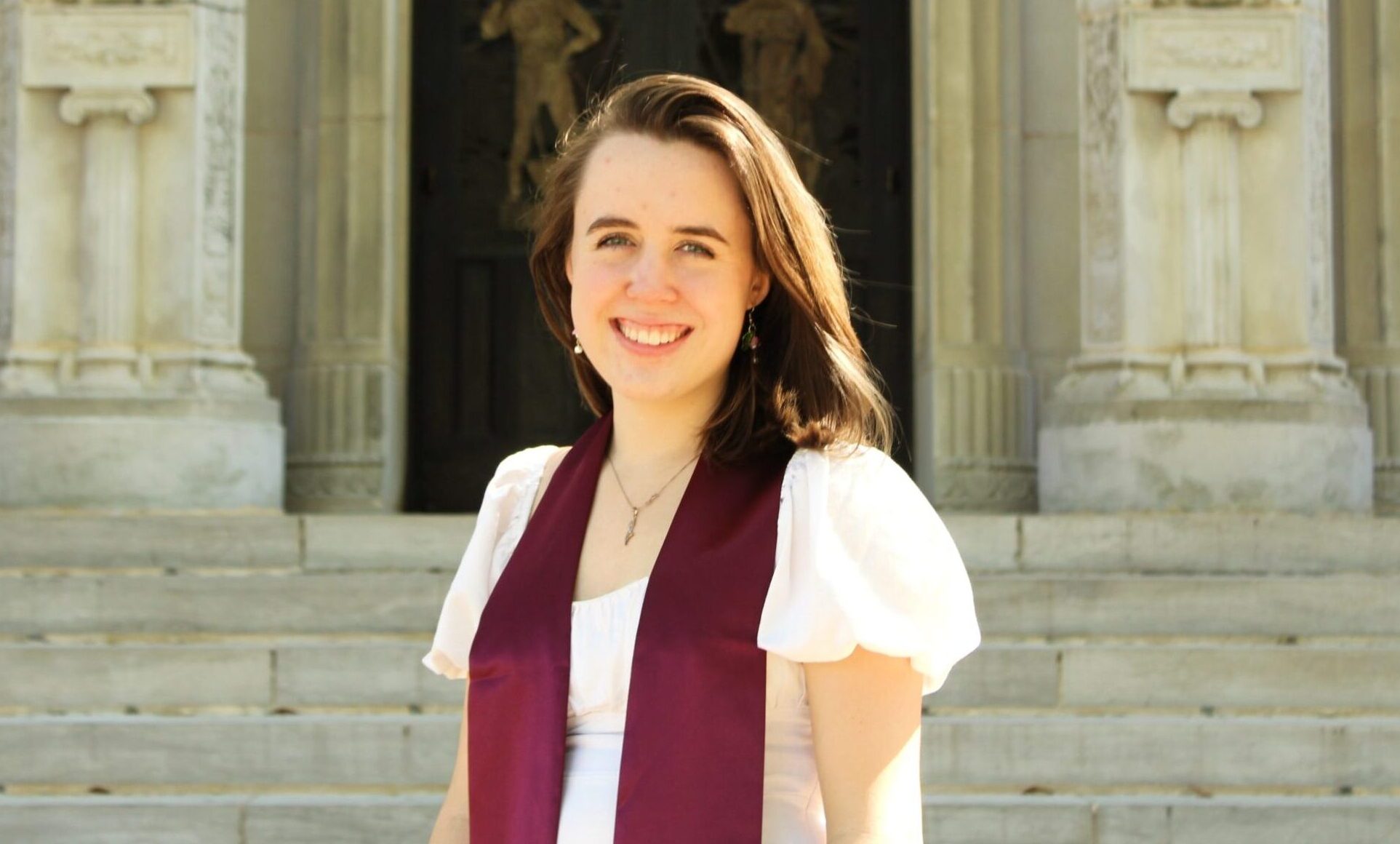 A college student wearing a white dress and maroon graduation sash stands outside of a university building as she smiles for a portrait.