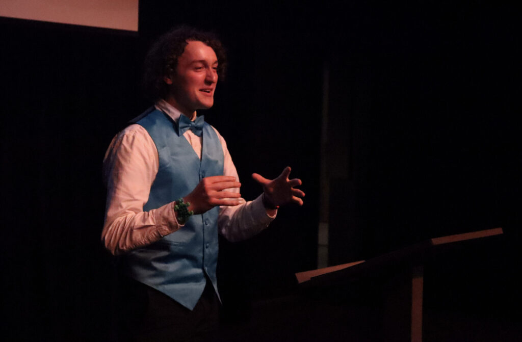 A college student wearing a vest and a bow tie performs onstage.