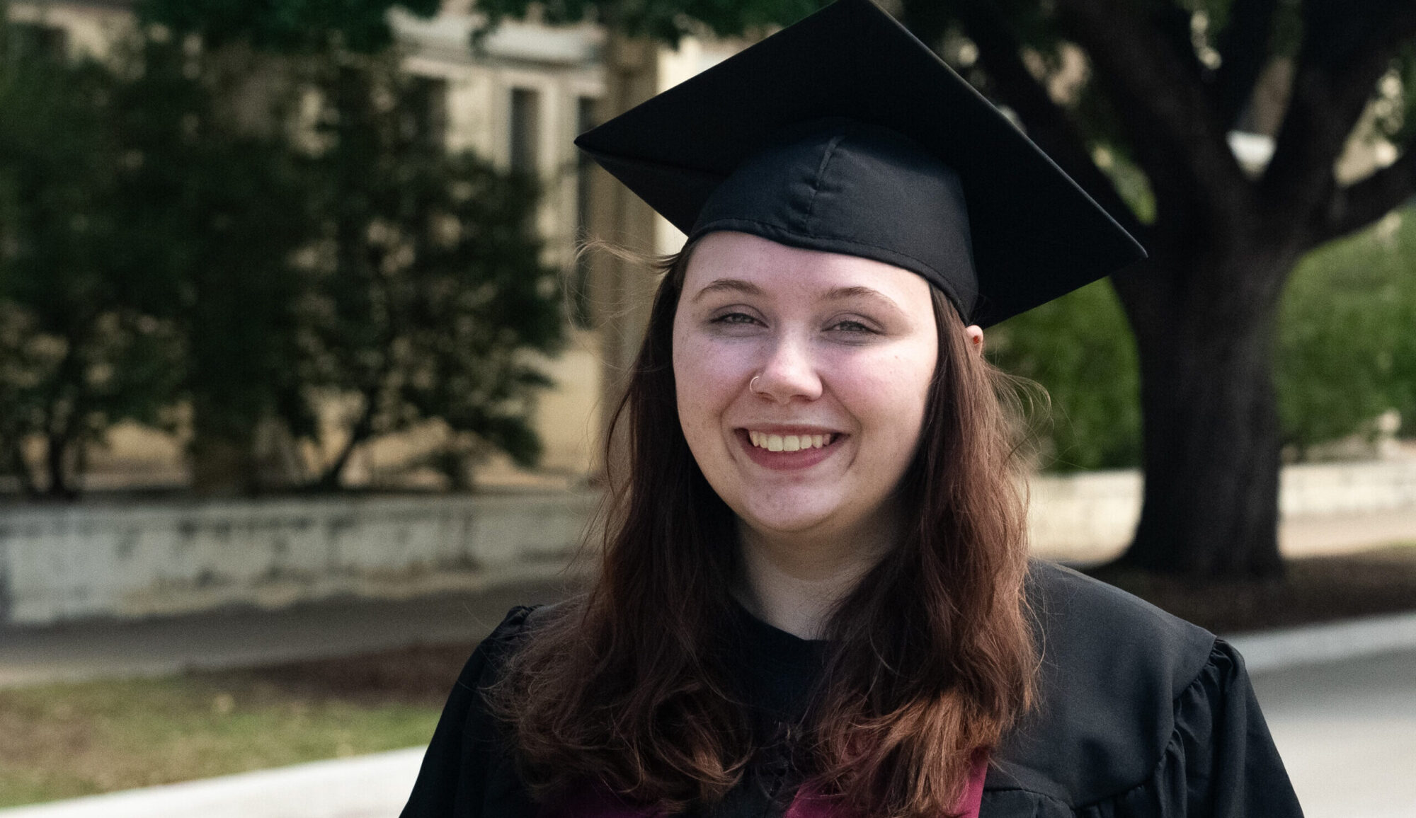 A college student wears a traditional graduation cap and gown and a maroon sash.