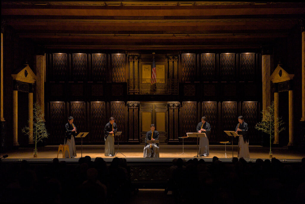 Five musicians, four of them standing, play shakuhachi music in a concert setting.