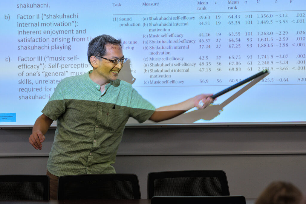 A shakuhachi player uses the bamboo flute to point to something projected on a screen behind him.