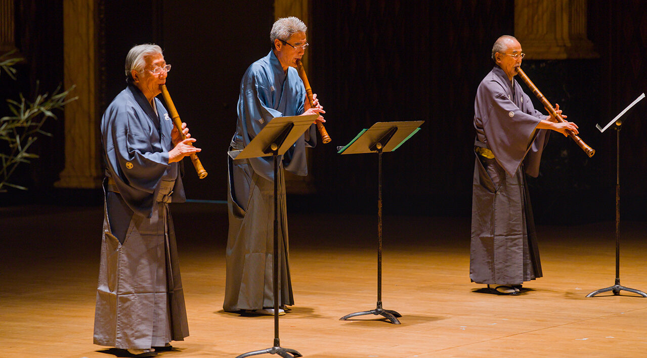 Three wearing robes perform with shakuhachi bamboo instruments on a stage.