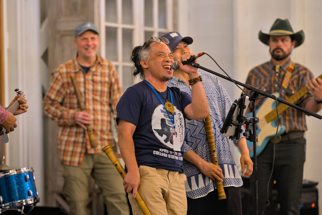 A man holding a microphone in his left hand and a shakuhachi bamboo flute in his right sings on stage, with other performers behind him.