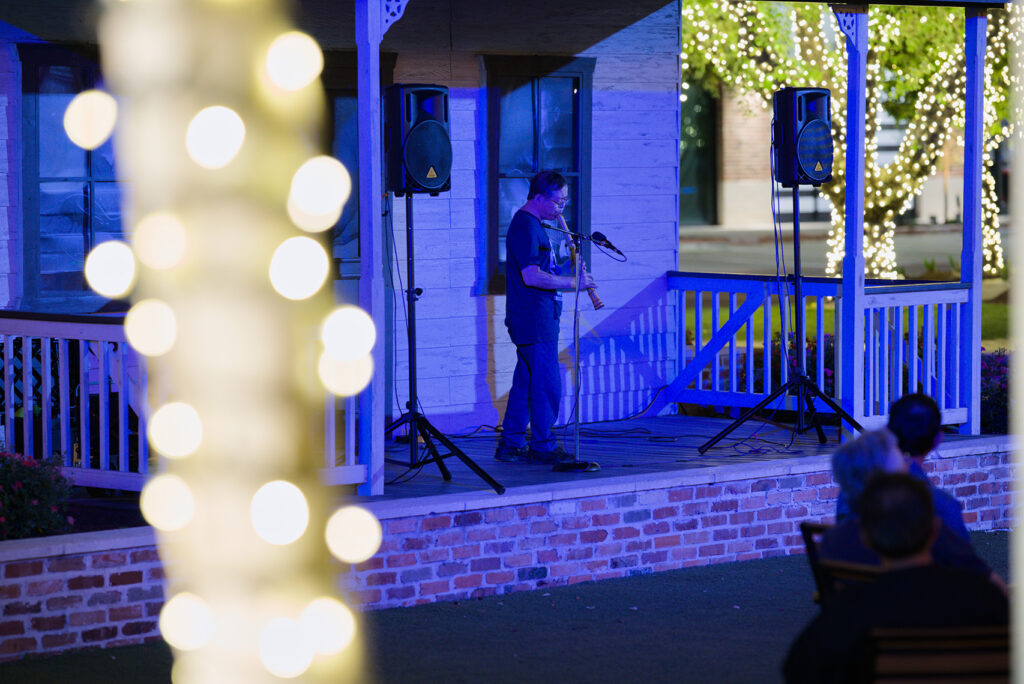 An artist performs with a shakuhachi bamboo flute in a nighttime concert on a porch structure.