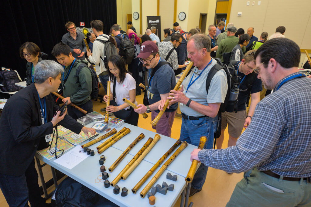 A crowded room of people looking at and holding shakuhachi bamboo instruments.