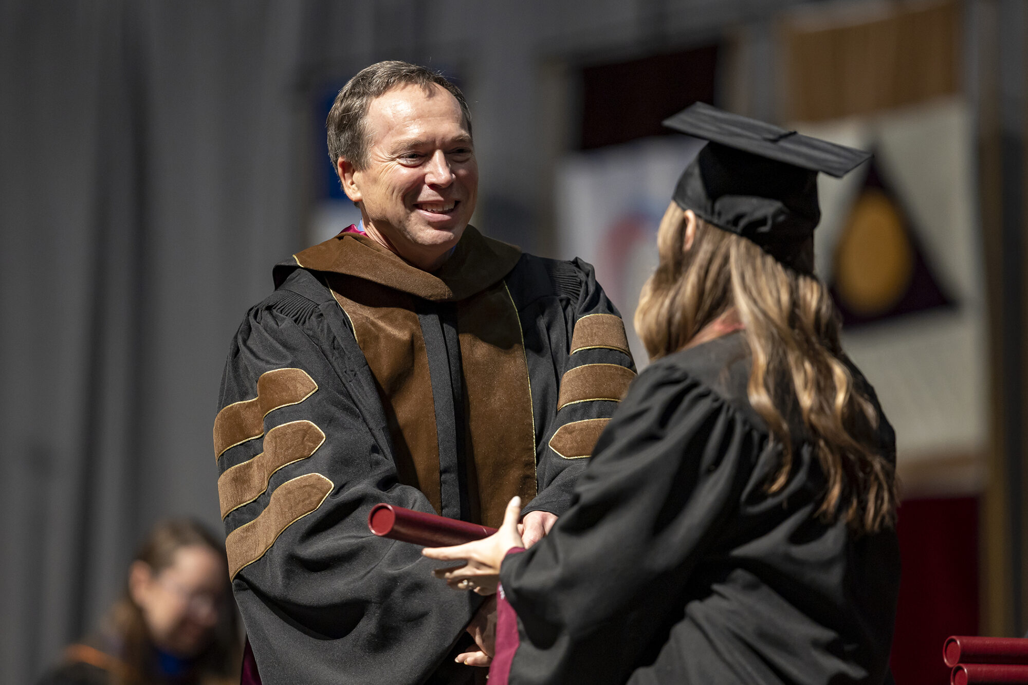 A school leader gives a diploma to a college student wearing a black cap and gown.