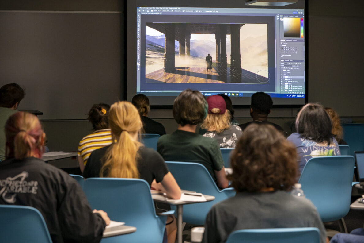 Students in a classroom look at a video game image being shown on a screen in front of them.