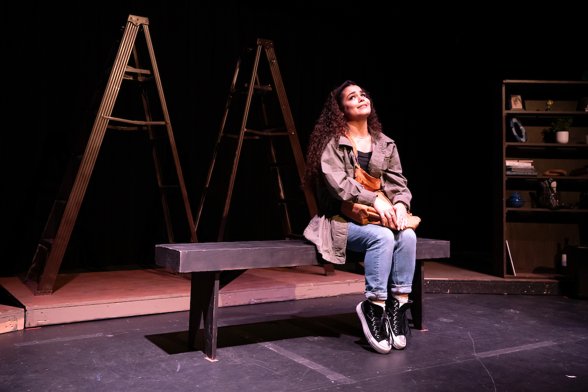 A student sits on a bench on a stage during a dress rehearsal. The main stage light shines down on her.