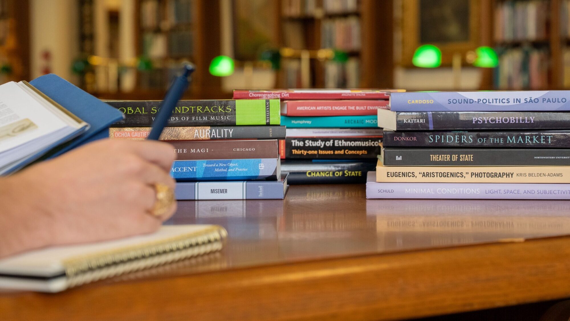 A hand with an Aggie ring takes notes near a stack of arts-themed books.