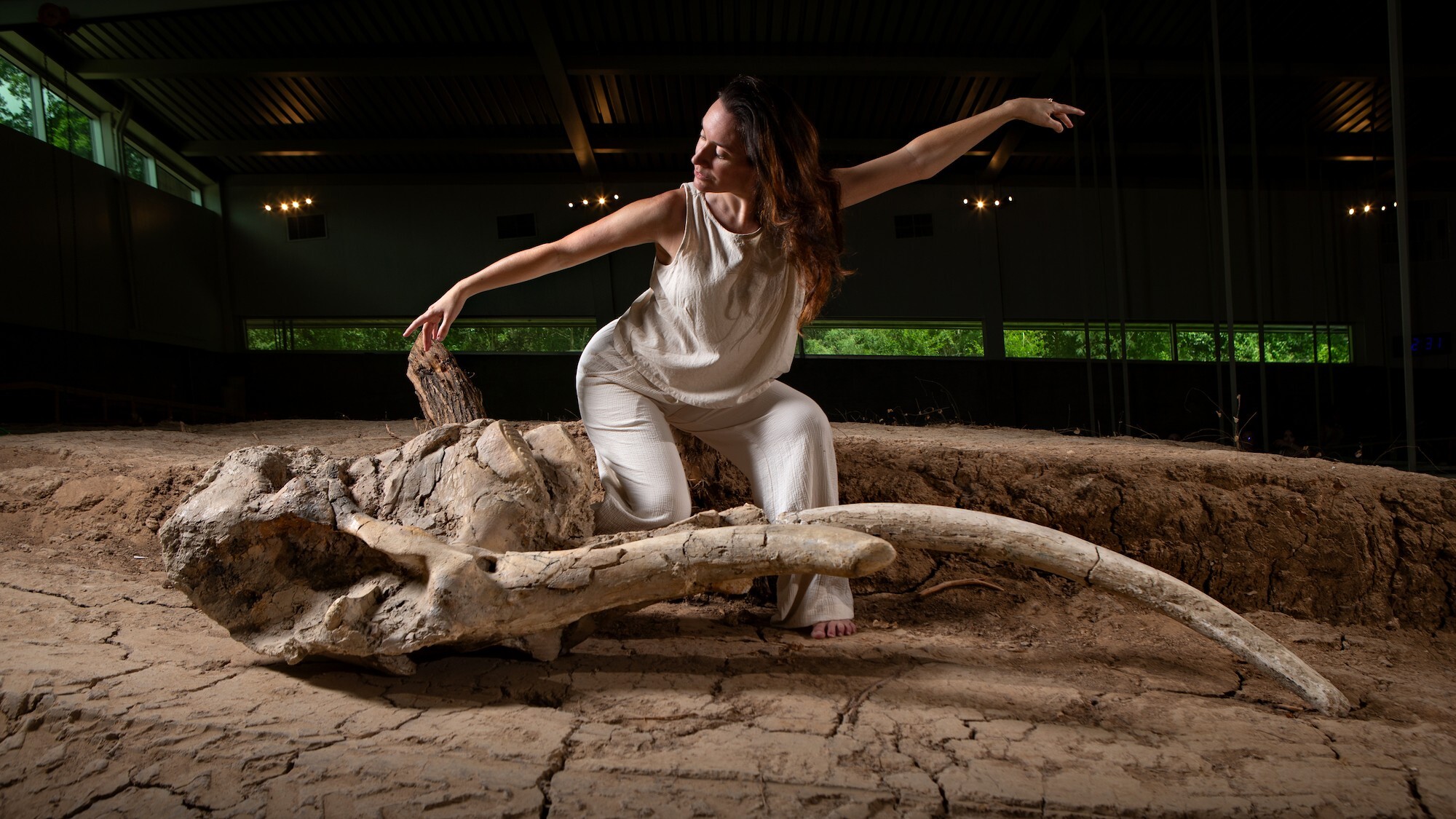 A university professor kneels with her hands out next to a giant mammoth fossil.