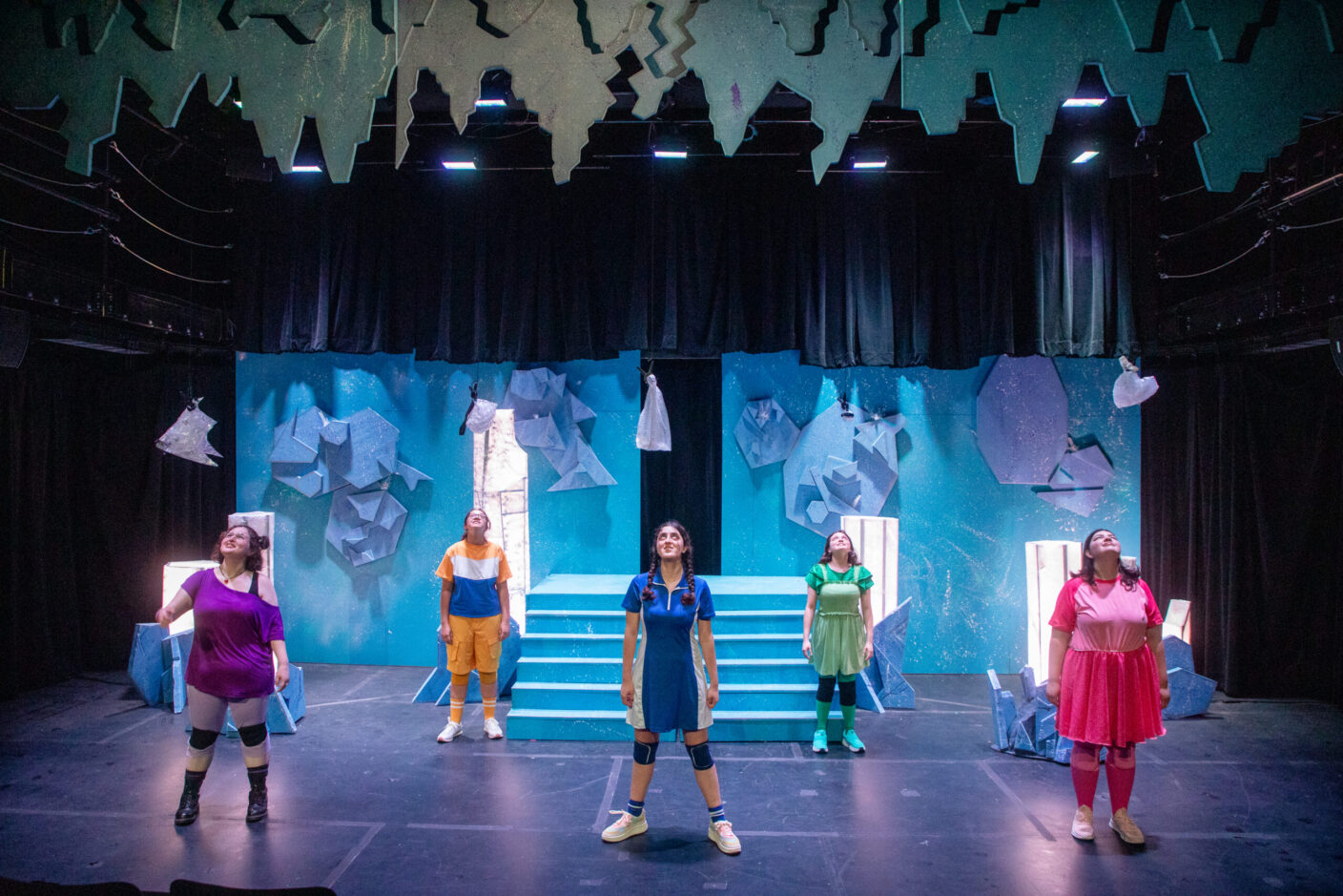 Five student actors, all dressed in a different color (purple, orange, blue, green and pink) perform on stage in a theatrical production.