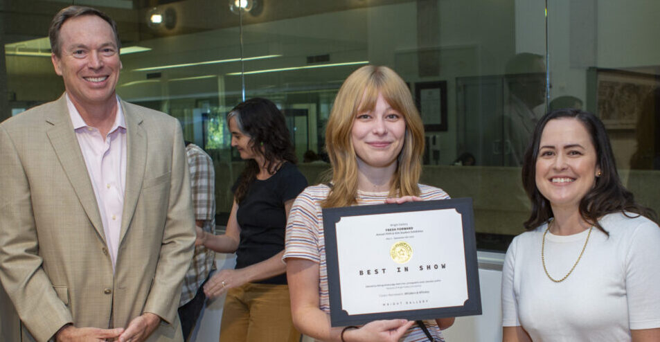 A university professor, student and other university professor stand in a gallery. The students holds a plaque that says "best in show."