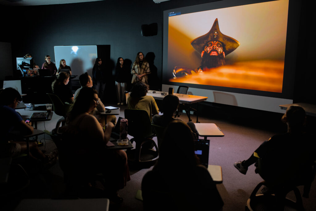A group of university students sit in a dimly lit classroom and watch a projection screen showing a pirate.