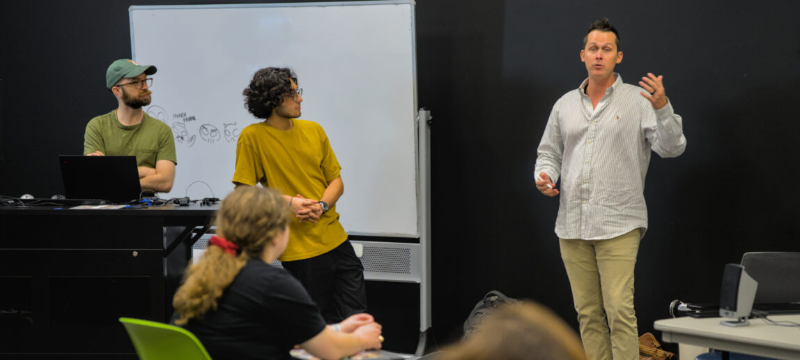 Three men stand in a university classroom.