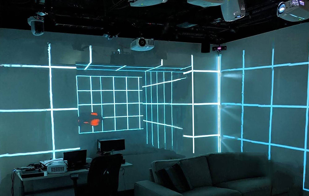 A photo of a dimly lit room with a couch, a table with electronics, and projector and light beams projected in square boxes on the wall.