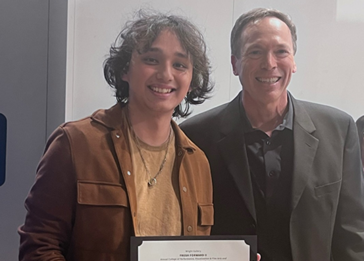 A university dean and a student stand in an art gallery surrounded by people. The student is holding a plaque award.