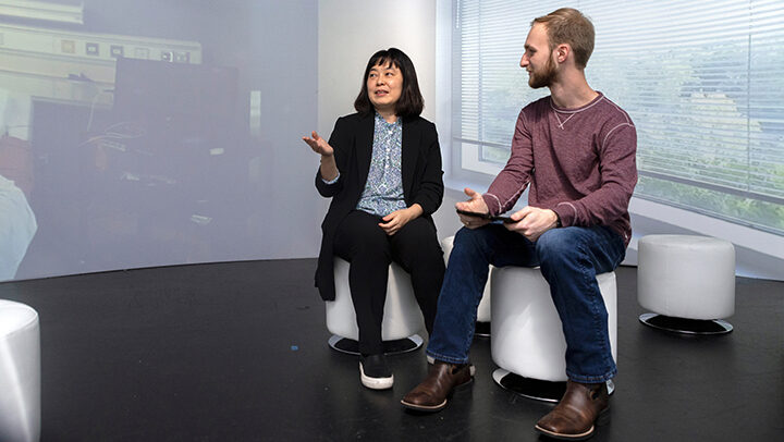A professor speaks with a student while seated inside a room with a 360-degree screen.