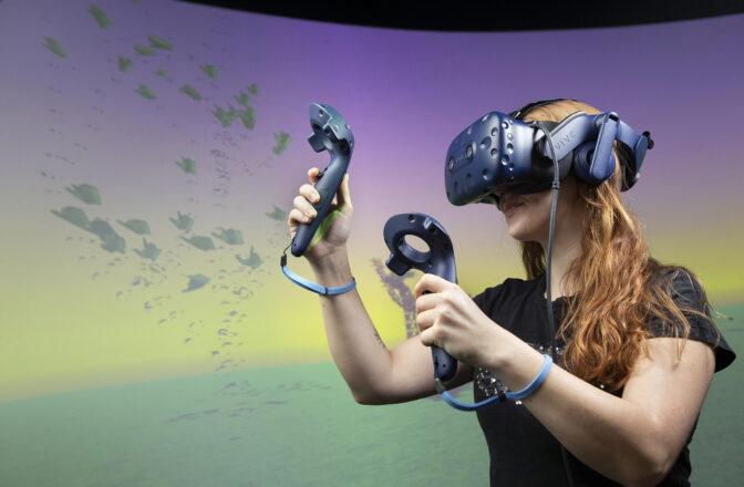 A woman wears a virtual reality headset and hand-held sensors in front of a colored background.