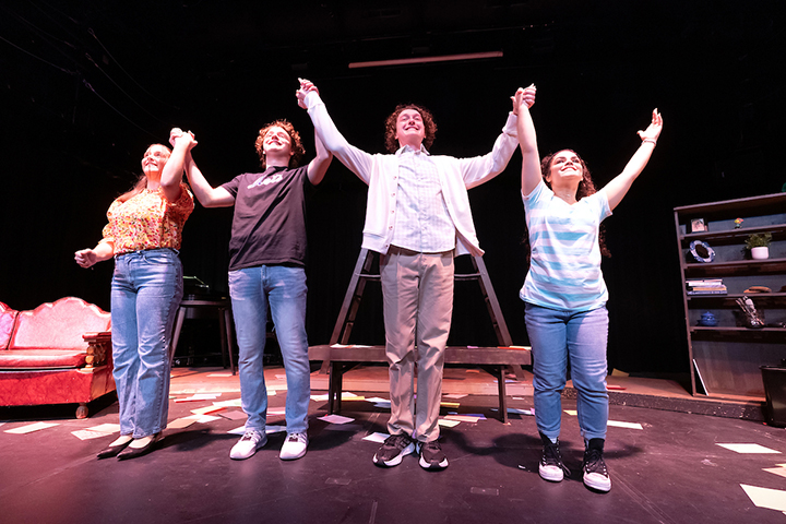Four college students raise their arms with joined hands at the end of a theater performance.