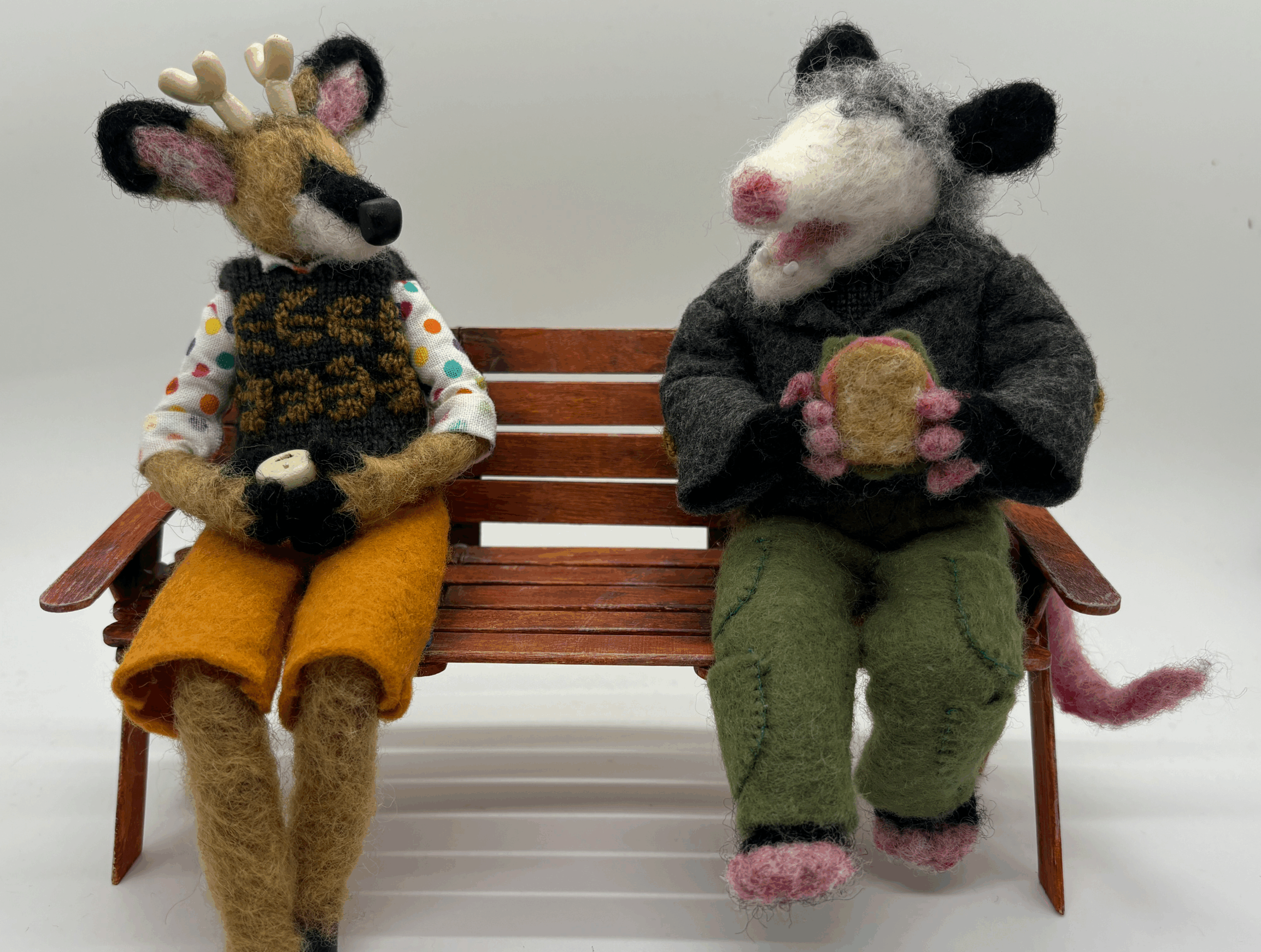Handmade felt sculptures of a deer and an opossum sitting on a wooden bench. They are sitting like people, wearing clothes and chatting with each other.