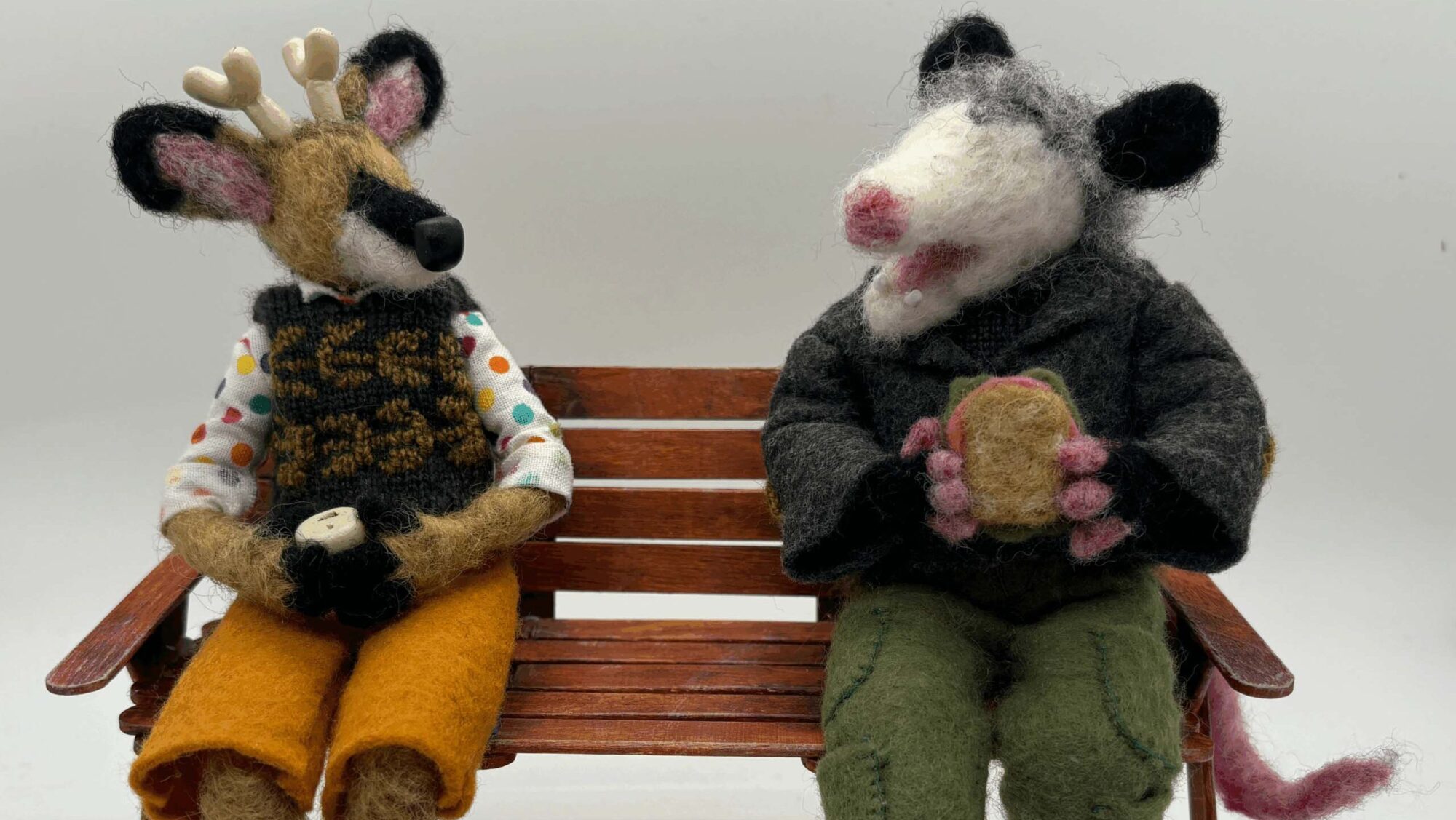 Handmade felt sculptures of a deer and an opossum sitting on a wooden bench. They are sitting like people, wearing clothes and chatting with each other.
