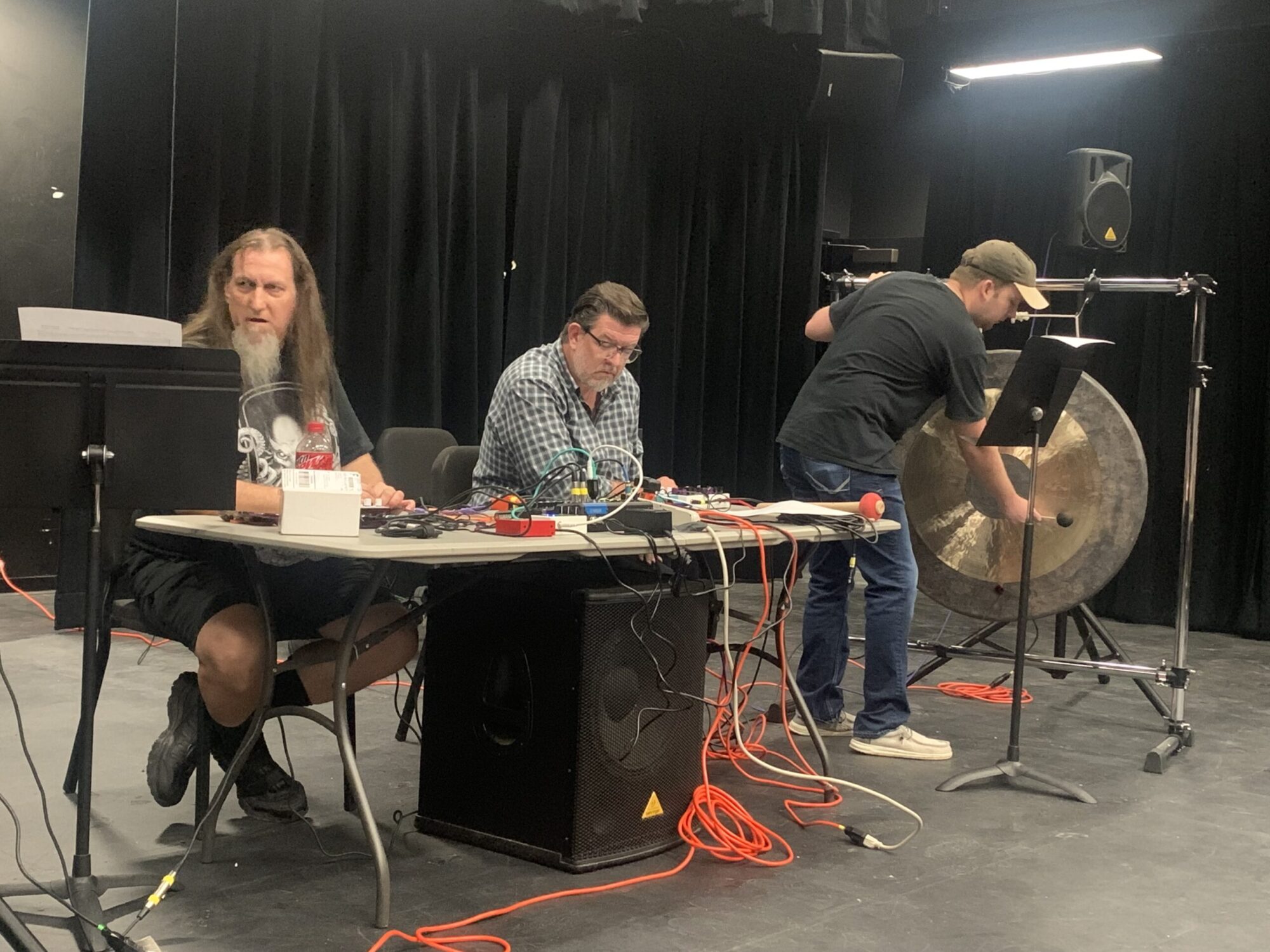 Two music professors are seated at a table, manipulating sounds with electronic equipment. To their left stands a college student playing the gong-like instrument known as the tam-tam.