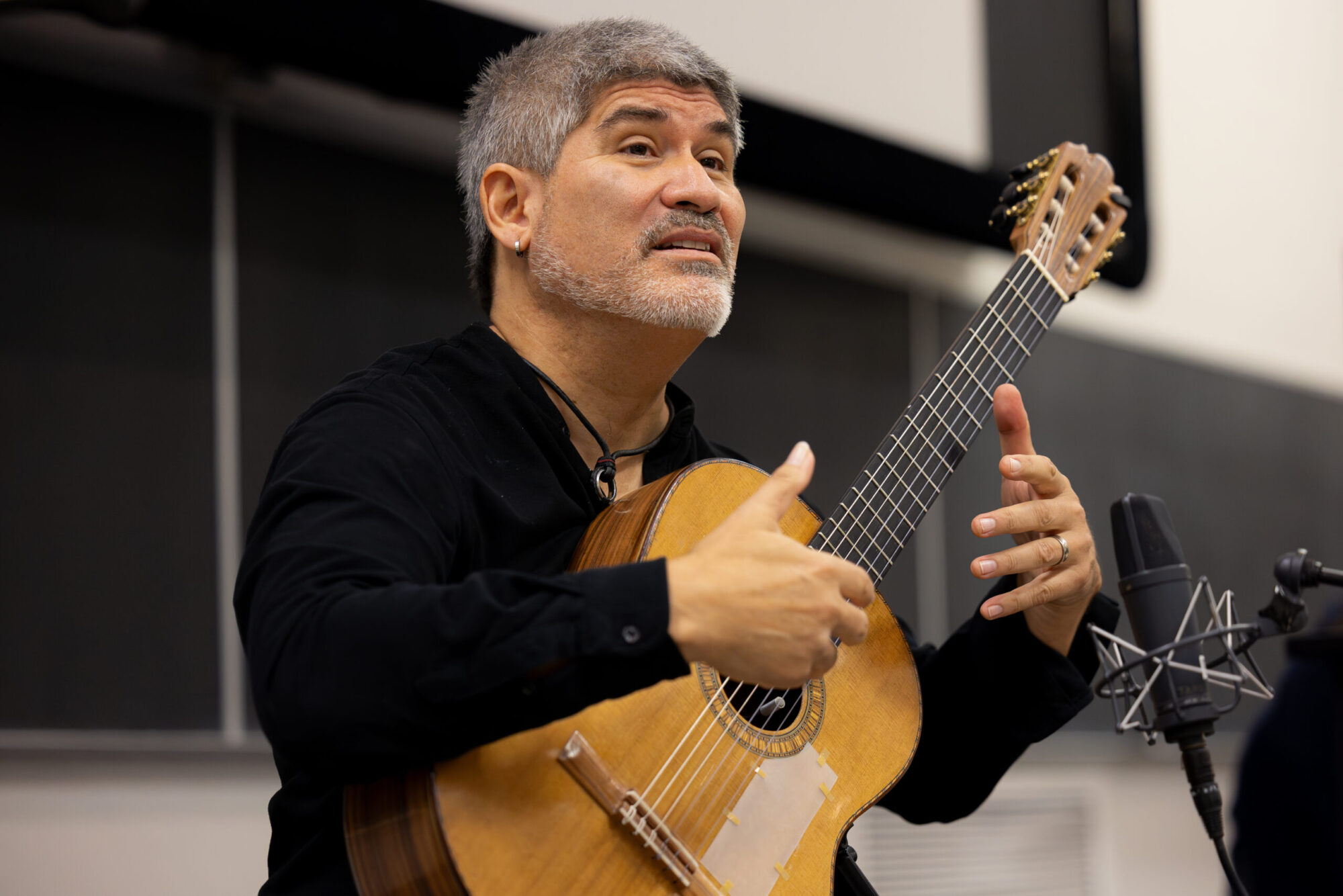 A man sits with a guitar while talking to college students in a classroom.