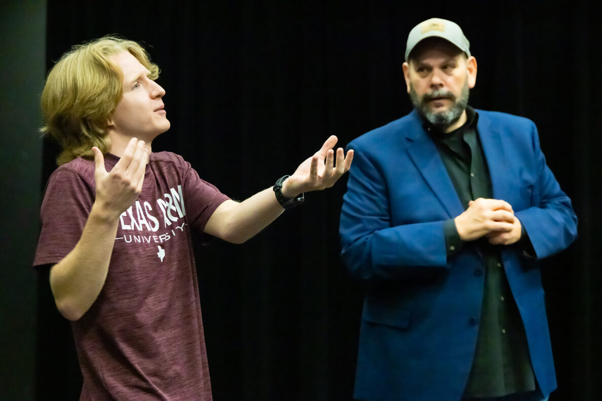 A college student stands in a theater at an improv-comedy workshop, speaking and gesturing his arms outward. A man stands to his left.