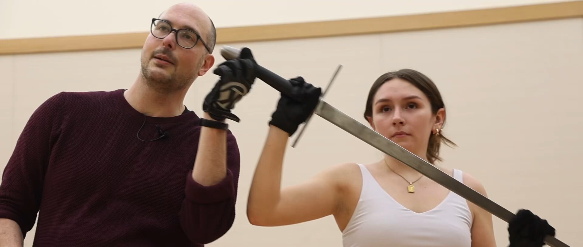 A college student holds a sword in a theatre training exercise, with her professor standing to her right.