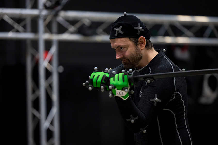 A man dressed in a bodysuit designed for motion capture holds a toy sword.