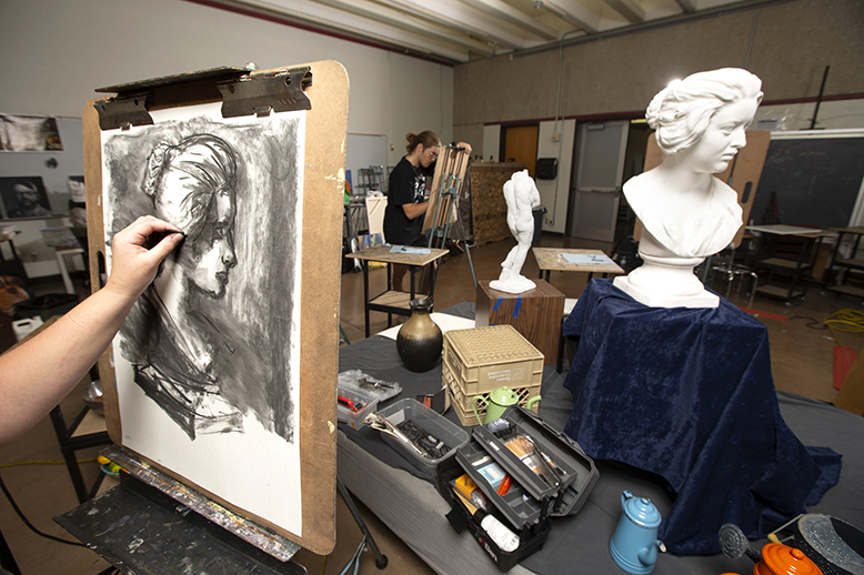 A college student works on a drawing while looking at the bust sculpture of a woman.