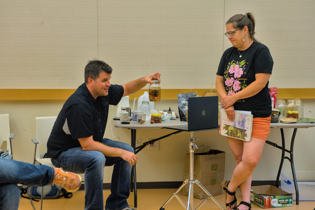 Two people in a classroom showcase various teas in glass jars. 