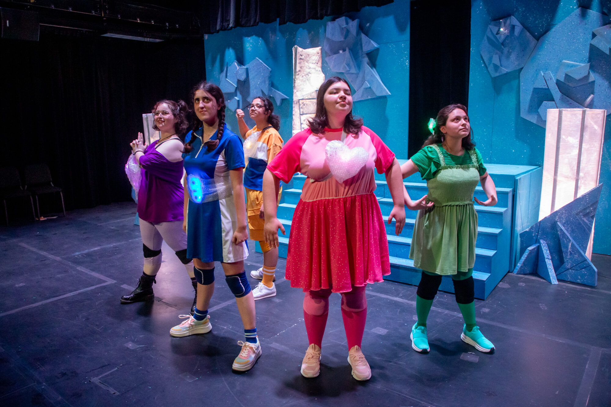 Five college students in colorful outfits pose on a stage during a theater performance.