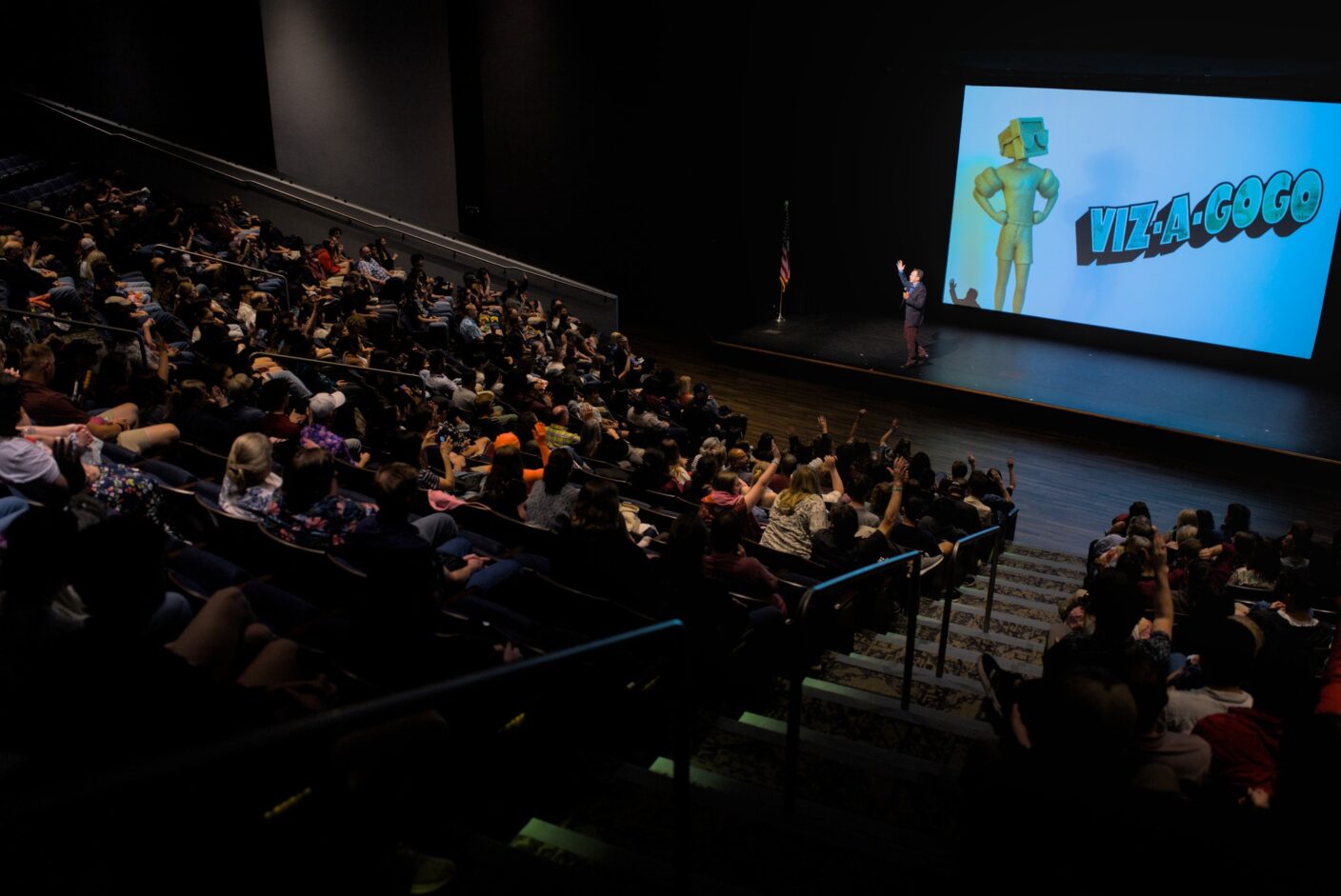A university dean stands before an audience in a theatre. On the screen behind him is an image that reads "Viz-a-GoGo 32."