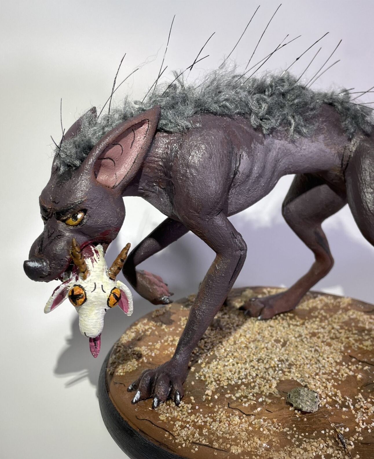 A handmade sculpture depicting a mostly hairless wolf-like creature holding a goat head in its snarling jaw. It has one paw lifted in mid-stride, and the hair it does have runs down the middle of its back, from head to tail. The pedestal it stands on is round, painted brown with a dusting of sand particulate across it.
