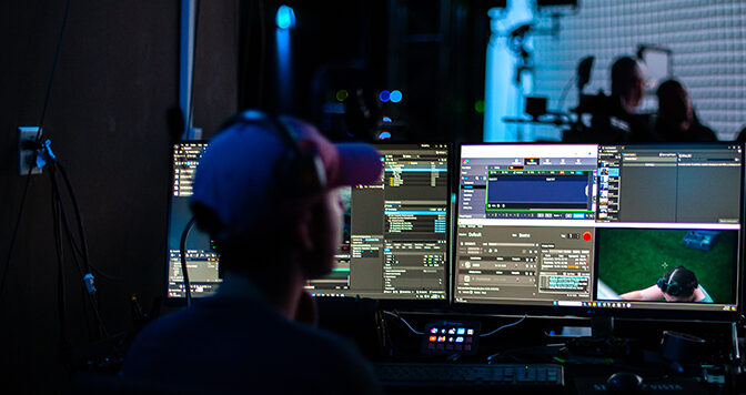 A student sits in front of multiple computer screens while a crew in the background works on a virtual production stage.