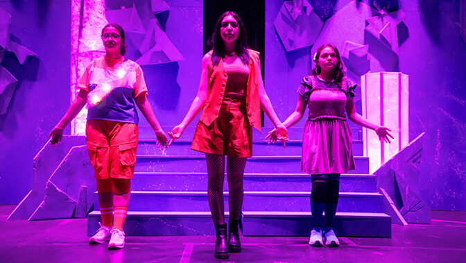 Students in bright costumes with a purple light overhead stand on a stage during a theatre production.