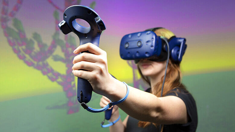 A college student wears a virtual reality headset and holds controllers in her hands, standing in front of a screen showing bright colors.