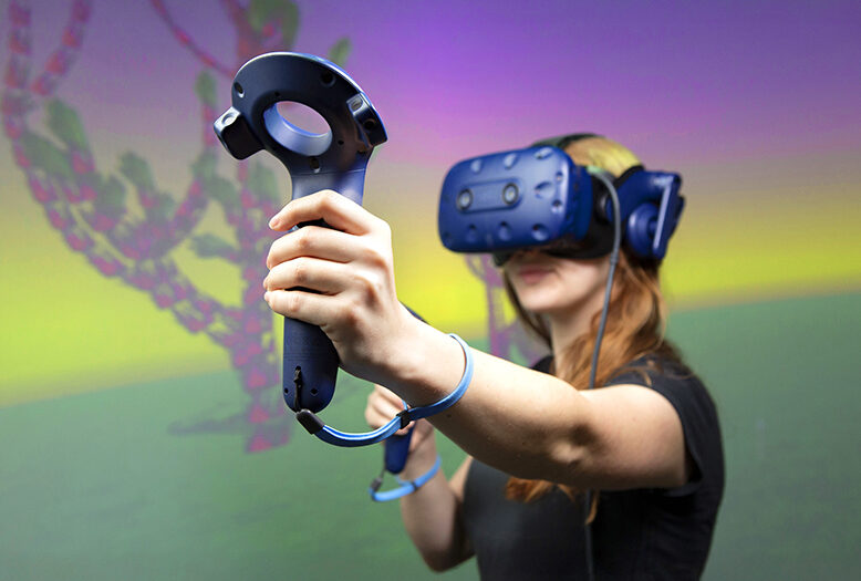A college student wears a virtual reality headset and holds controllers in her hands, standing in front of a screen showing bright colors.