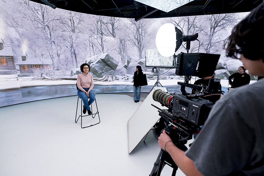 A student sits in a chair in front of a large LED wall showing a winter scene on a virtual production stage.