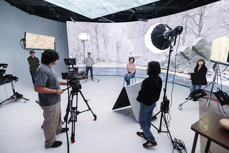 A student sits in a chair in front of a large LED wall showing a winter scene on a virtual production stage.