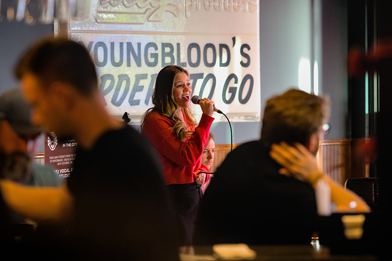 A college student sings into a microphone in a busy bar setting.