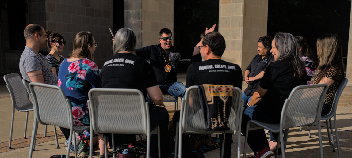 People sit in chairs outside a university courtyard and look at a drum, one man has his hands out and is holding a drum stick.