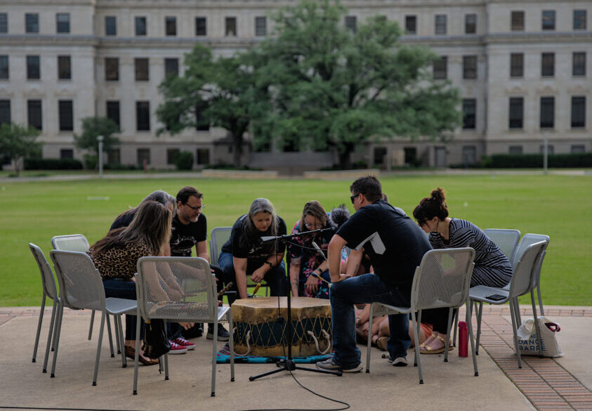 People sit in chairs outside a university courtyard and look at a drum, one man has his hands out and is holding a drum stick.