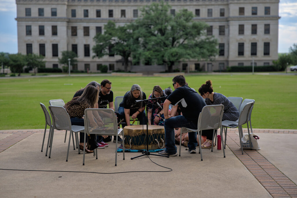 People sit in chairs outside a university courtyard and look at a drum, one man has his hands out and is holding a drum stick.