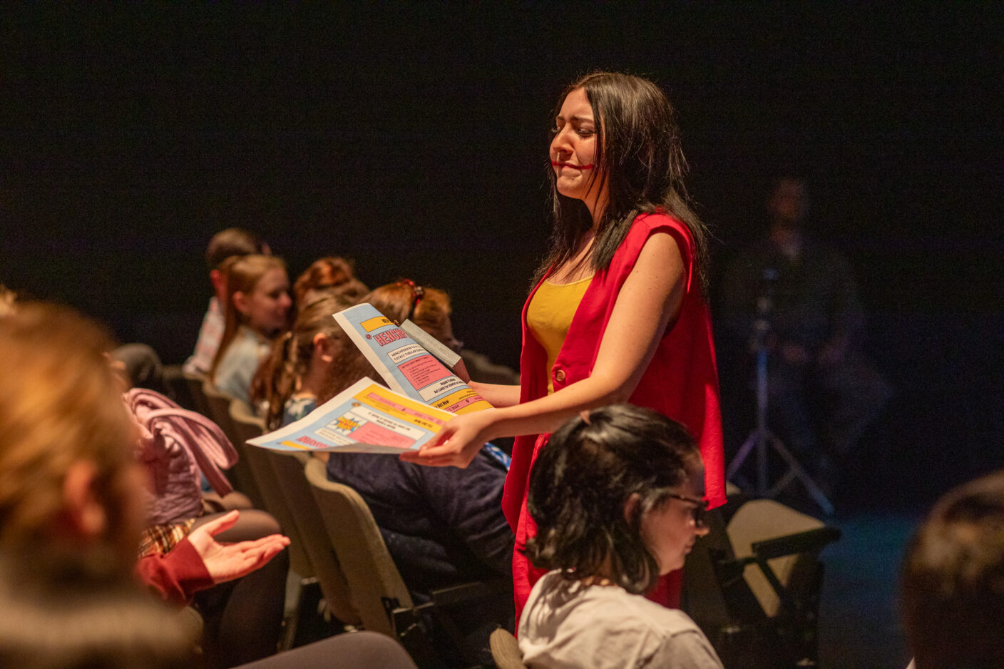 A college student acting in a play goes into the audience and hands out fliers.