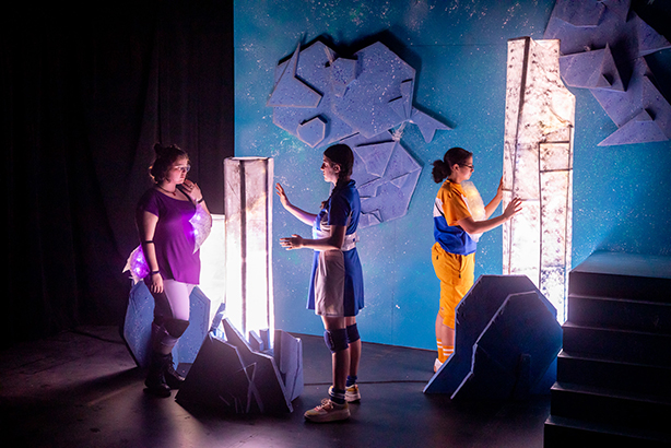 Students in bright costumes stand next to bright light structures in a theatre production.